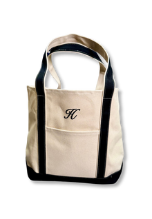 Custom Tote, Superior MI Tote, XL, Open Top, Helmsman Stitch Company. A cream canvas tote bag with black trim and dual shoulder straps, featuring a front pocket with an embroidered black monogram letter 'H' on the front, set against a plain white background. Makes great gifts.