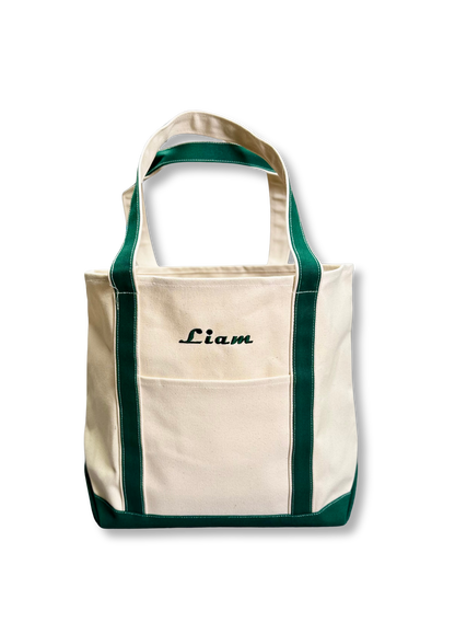 Custom Tote, Superior MI Tote, XL, Open Top, Helmsman Stitch Company. An off-white canvas tote bag with dark green trim and handles, featuring a front pocket and the name 'Liam' embroidered in black script on the upper front. Makes a great gift.