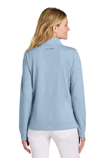 Woman wearing a light blue jacket with a visible brand logo on the back, standing against a white background. TravisMathew Women's Coveside 1/2-Zip, Embroidered