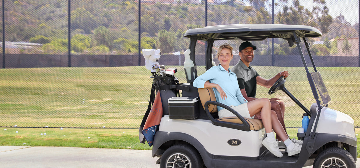 A woman in a light blue polo shirt and a man in a dark polo shirt sit in a white and gray golf cart on a golf course, with a golf bag loaded with clubs behind them. TravisMathew Women’s Onward 1/2-Zip, Embroidered