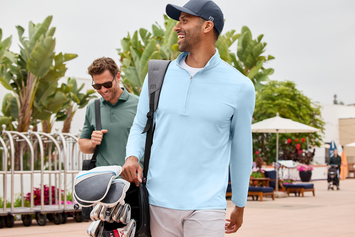 Two men walking outdoors with golf bags, one wearing a light blue shirt and cap. TravisMathew Onward 1/2-Zip, Embroidered