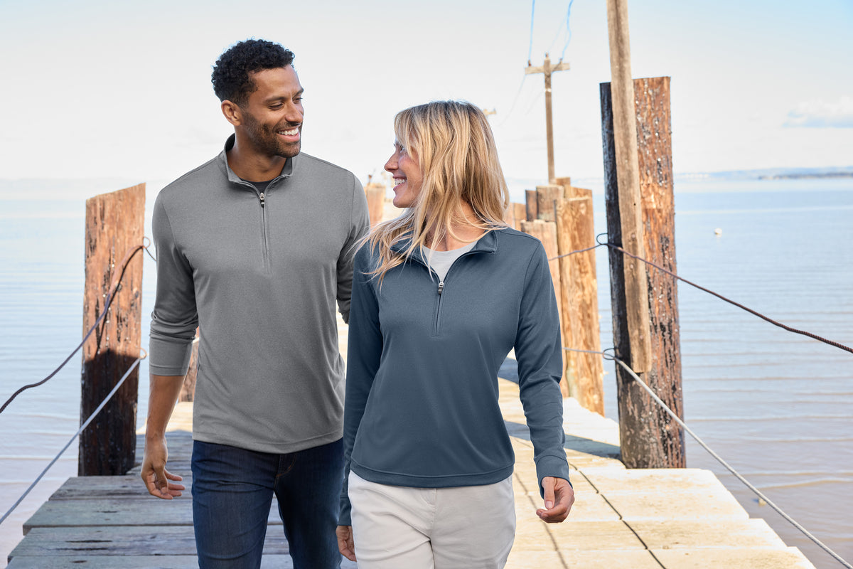 A man and a woman walking side by side on a wooden dock by the water, wearing matching long-sleeve quarter-zip pullovers in gray and navy blue. TravisMathew Women's Coto Performance 1/2-Zip, Embroidered