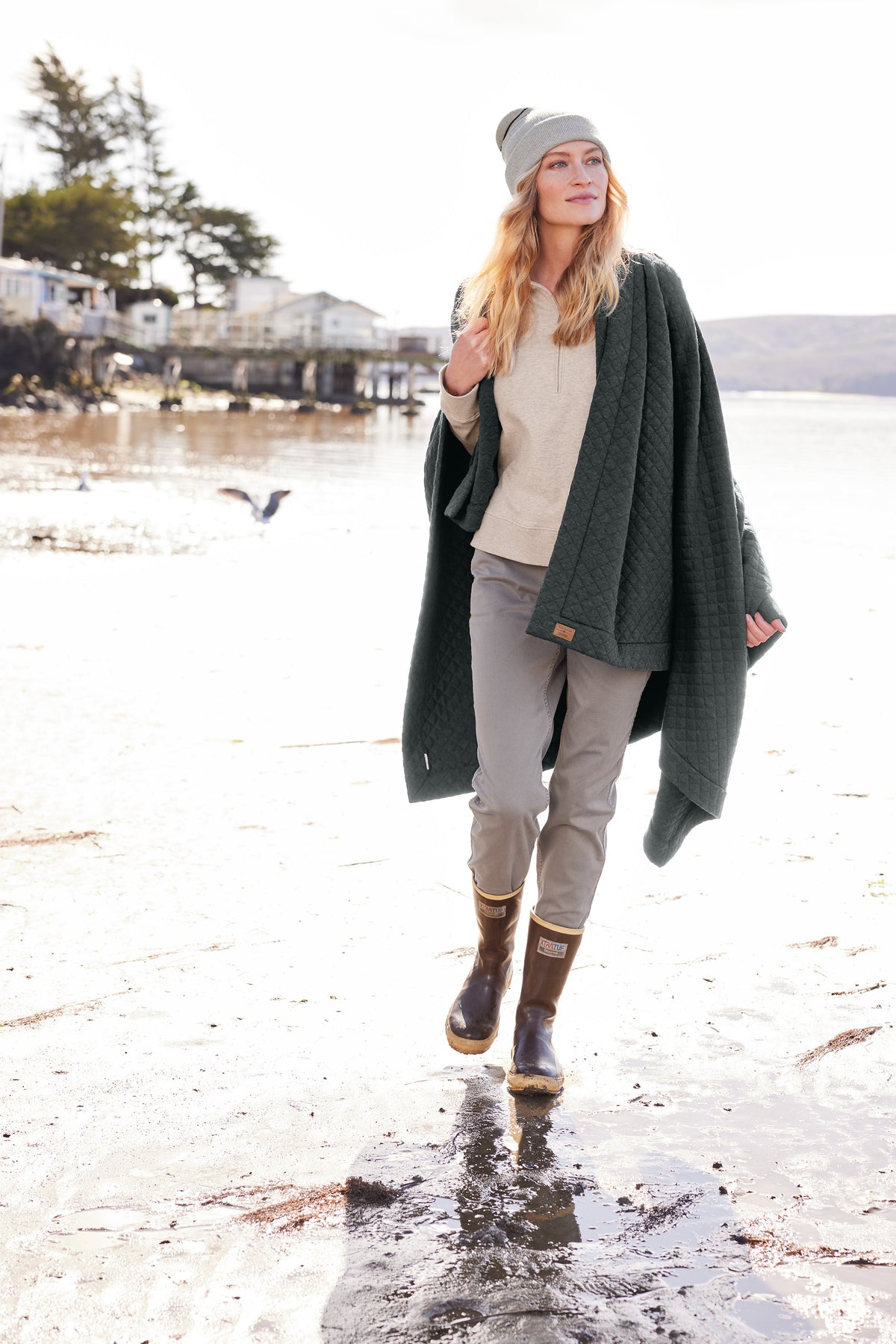 Woman walking on a beach wearing a green coat, beige sweater, gray pants, and rain boots. TravisMathew Women's Ocean Villa 1/4-Zip, Embroidered