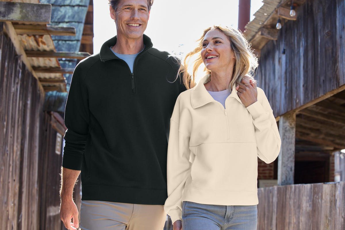 A man and a woman walking side by side outdoors between rustic wooden buildings. The man wears a black quarter-zip pullover over a light blue shirt, and the woman wears a cream quarter-zip pullover over a light purple top. TravisMathew Momentum Shift 1/4-Zip, Embroidered