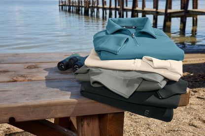A stack of folded quarter-zip polo shirts in teal, cream, sage green, and black sits on a wooden dock bench with water and a pier in the background. TravisMathew Momentum Shift 1/4-Zip, Embroidered