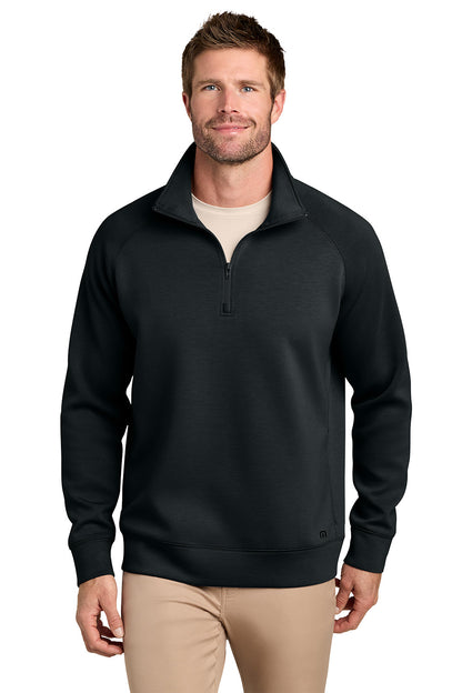 A man wearing a black quarter-zip pullover sweater with a stand collar, paired with beige pants, photographed against a plain white background. TravisMathew Momentum Shift 1/4-Zip, Embroidered