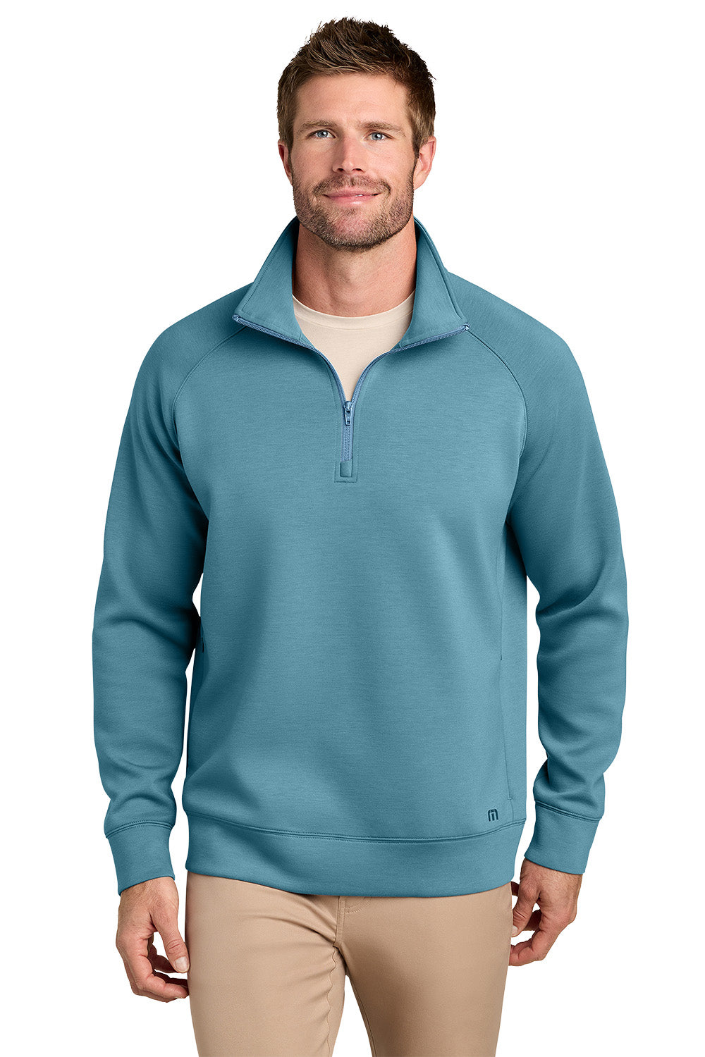 A man wearing a teal blue quarter-zip pullover sweater over a beige undershirt and khaki pants, photographed against a solid white background. TravisMathew Momentum Shift 1/4-Zip, Embroidered