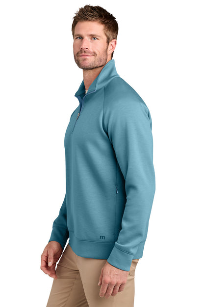 A man wearing a teal blue quarter-zip pullover sweatshirt and khaki pants, shown from a side profile against a white background. TravisMathew Momentum Shift 1/4-Zip, Embroidered