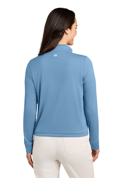 A woman shown from the back wearing a light blue long-sleeve quarter-zip pullover with a small white logo on the upper back, paired with off-white pants, against a plain white background. TravisMathew Women's Coto Performance 1/2-Zip, Embroidered