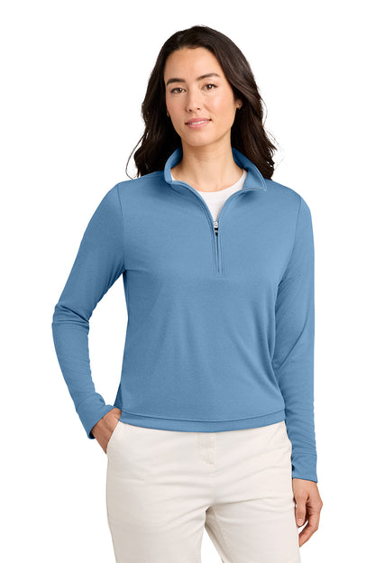A woman modeling a light blue long-sleeve quarter-zip pullover with a stand collar, paired with off-white pants, against a plain white background. TravisMathew Women's Coto Performance 1/2-Zip, Embroidered