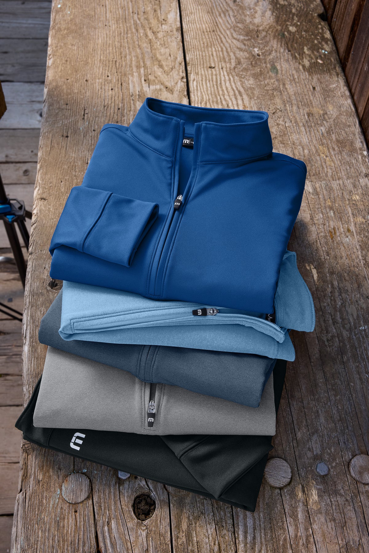 A stack of folded quarter-zip performance jackets in blue, light blue, gray, and black, arranged on a rustic wooden surface. TravisMathew Women's Coto Performance 1/2-Zip, Embroidered