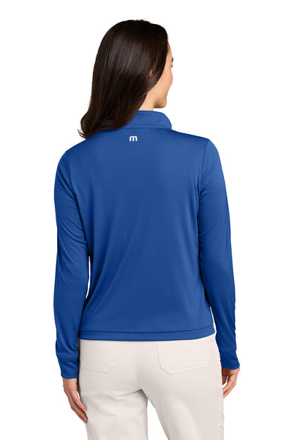 A woman viewed from the back wearing a royal blue long-sleeve quarter-zip pullover with a small white logo on the upper back, paired with light beige pants, against a plain white background. TravisMathew Women's Coto Performance 1/2-Zip, Embroidered