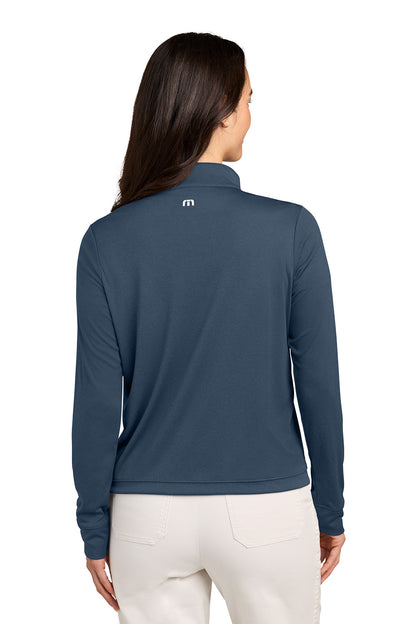 A woman viewed from the back wearing a slate blue long-sleeve quarter-zip pullover with a small white logo on the upper back, paired with off-white pants, against a plain white background. TravisMathew Women's Coto Performance 1/2-Zip, Embroidered